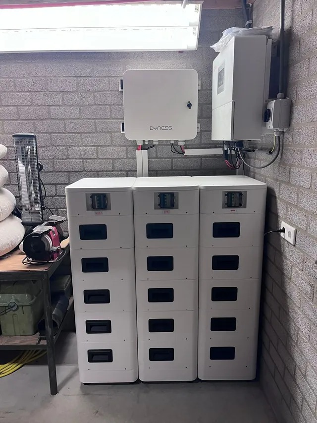 Drie witte Dyness-batterijtorens naast elkaar tegen een bakstenen muur, met daarboven een Dyness-aansluitkast en een Solis-omvormer.