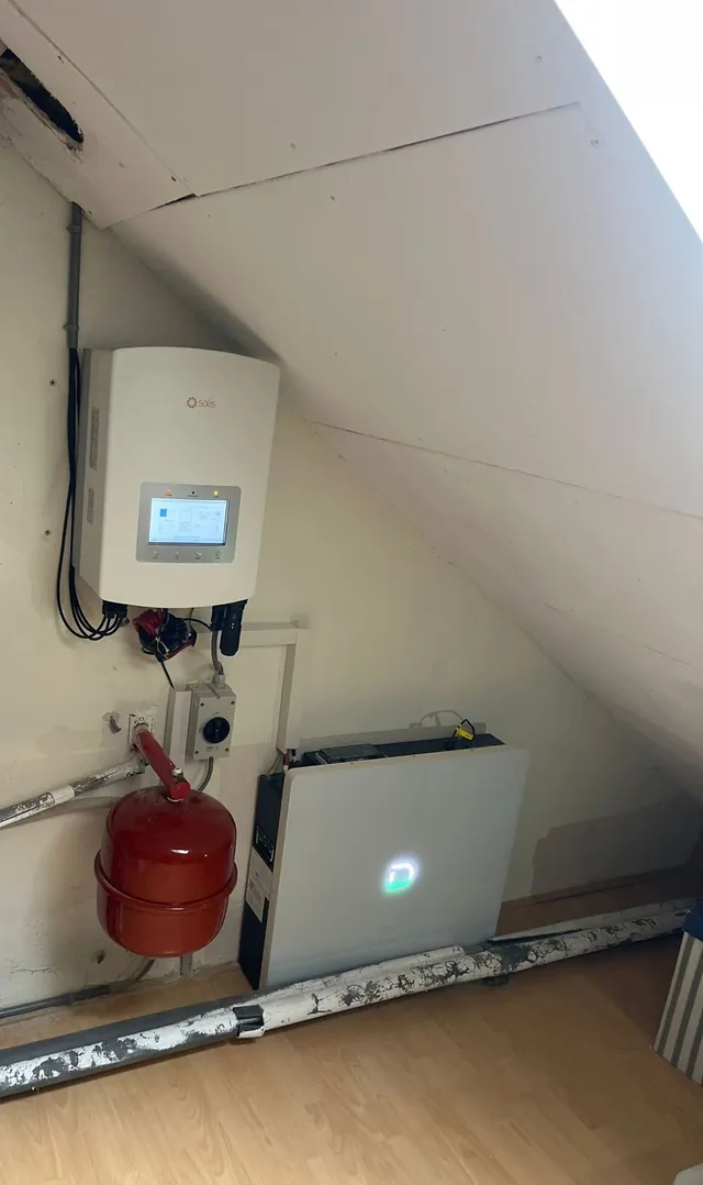 Compacte opstelling op zolder: een Solis-omvormer aan de wand en een platte Dyness-batterij op de vloer onder een schuin dak.